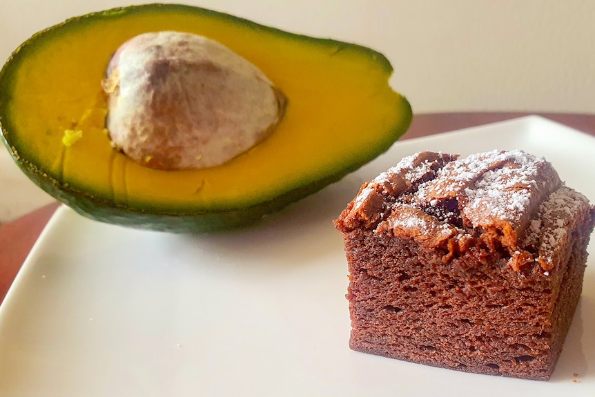 An avocado sits behind a chocolate brownie.