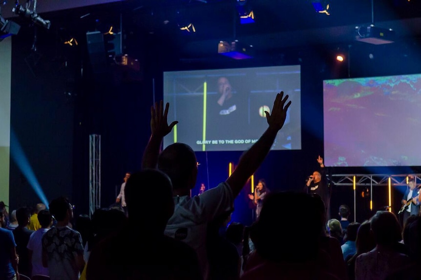 Worshippers raise their arms while a man speaks into a microphone on stage.