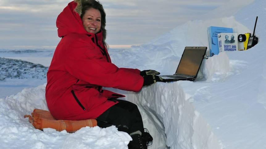 A woman sits on a pillow in the ice of Antarctica, dressed in a bright puffy jacket working on a laptop resting on the snow.