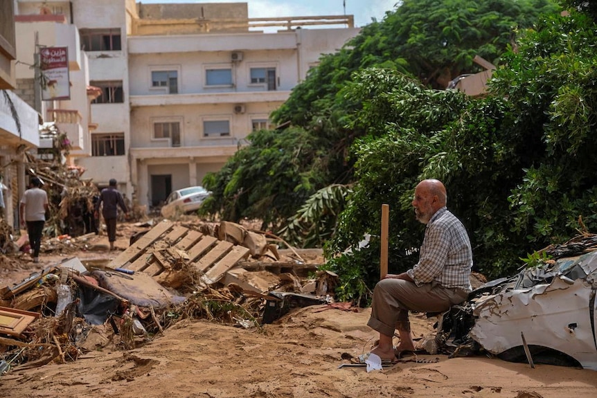 An elderly man sits on a damaged car surrounded by flood debris, multi-story buildings in the background