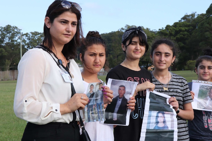 Five young women with dark hair stand in a line holding photos of people with grass and trees in the background.