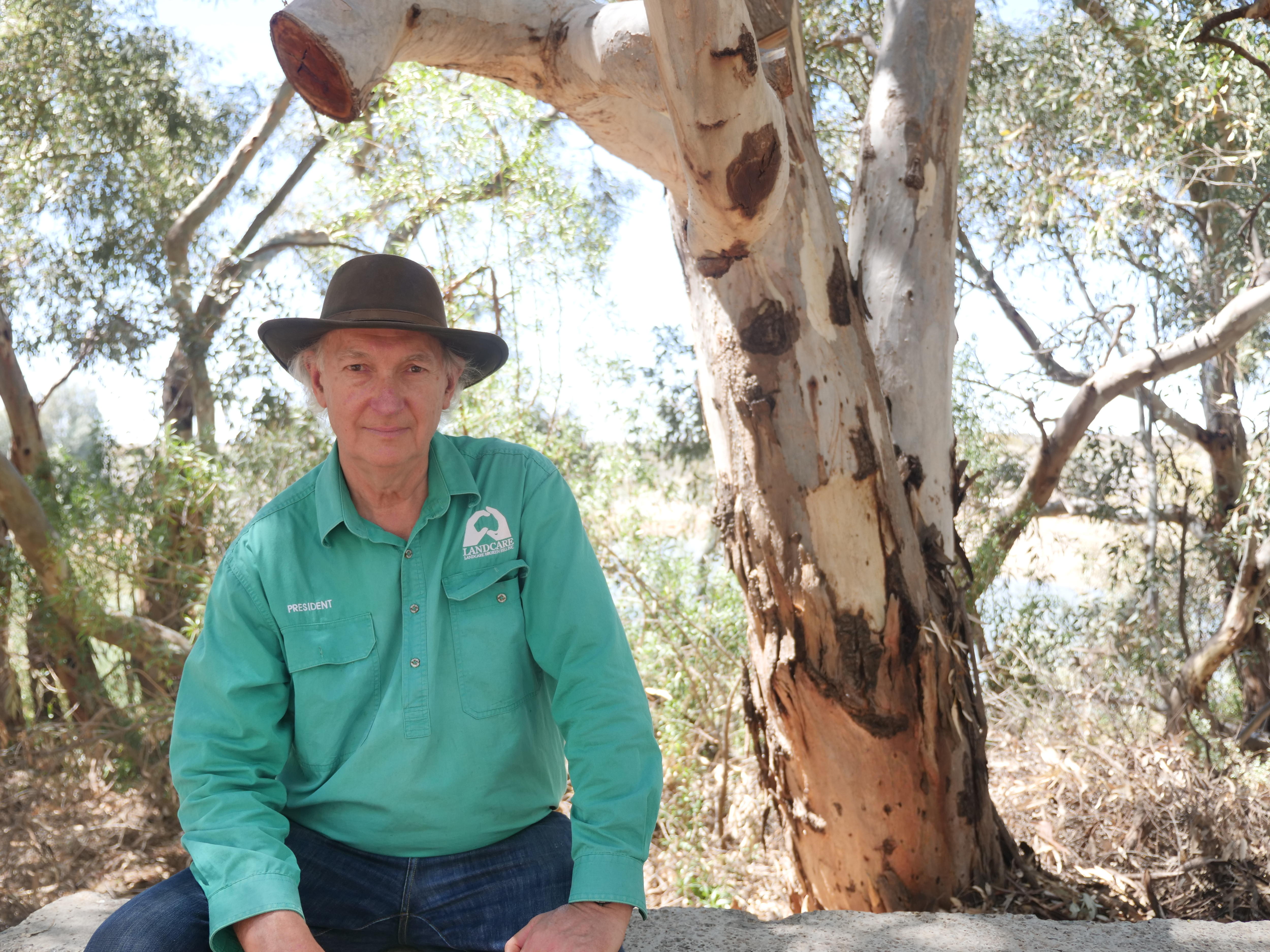 Simon veste uma camisa Landcare de mangas compridas e um chapéu de abas largas, sentado em um banco em frente a uma árvore no Parque Natural.