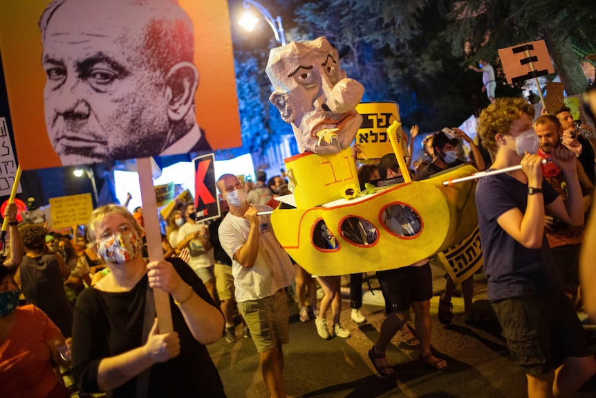 Demonstrators wearing face masks carry a float and a banner mocking Israel's Prime Minister Benjamin Netanyahu