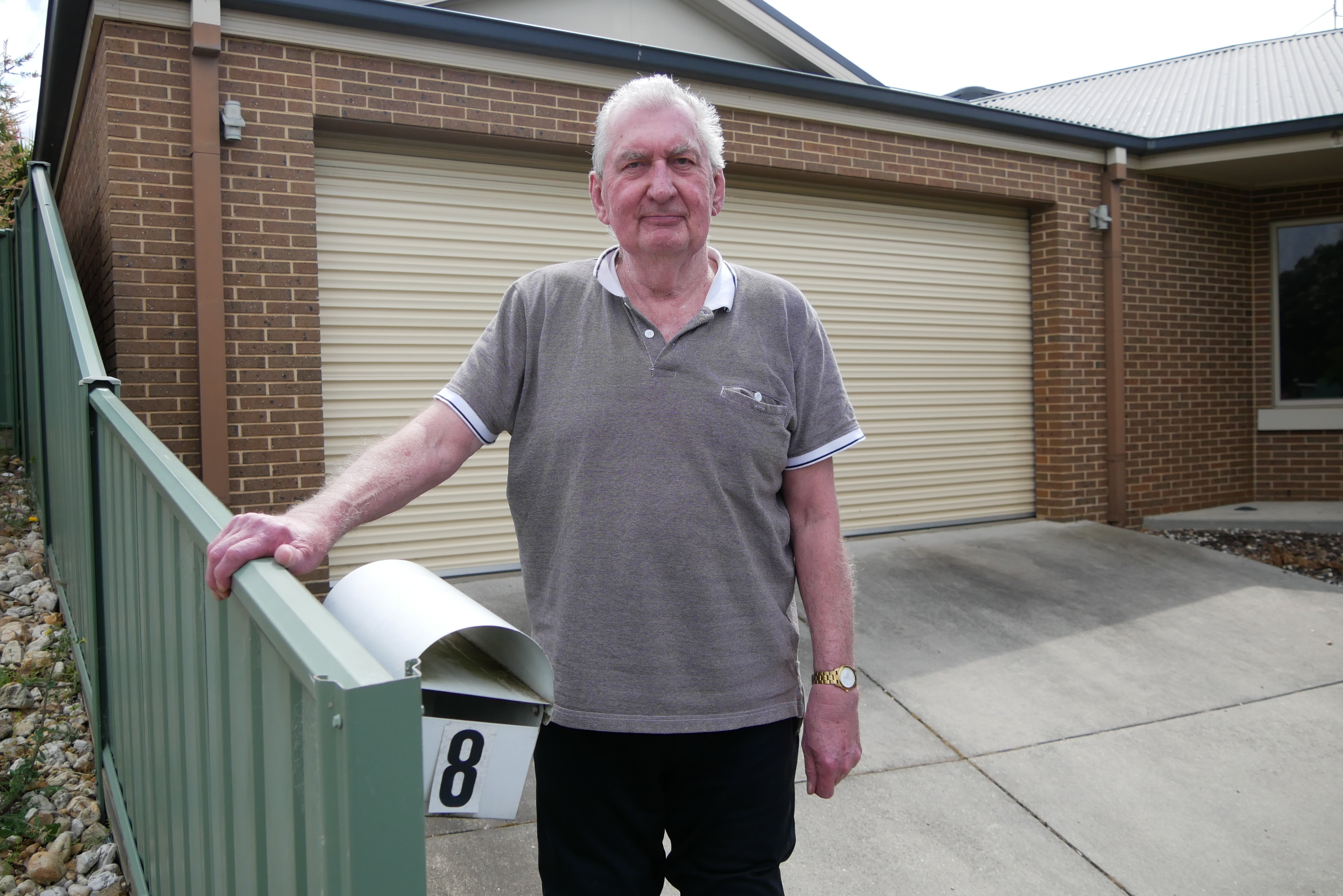A man in a grey shirt stands in front of a close carport in a driveway