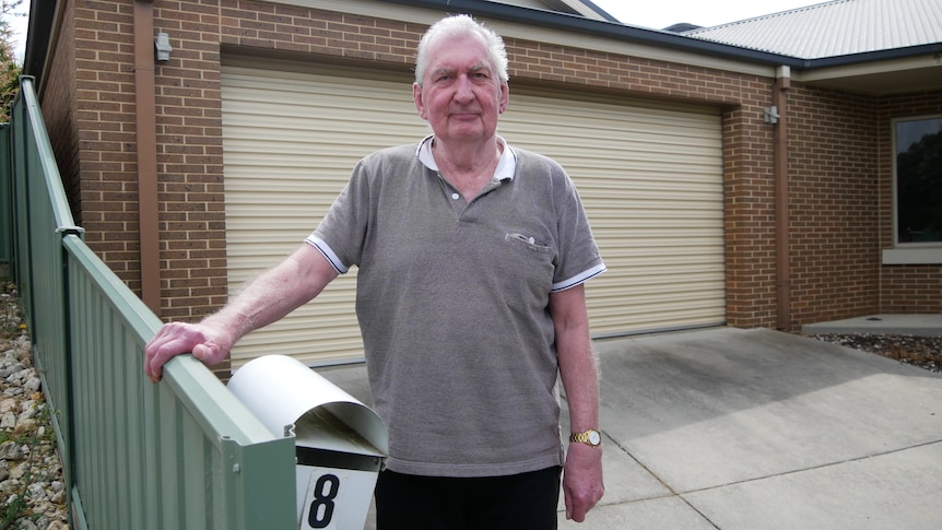 A man in a grey shirt stands in front of a close carport in a driveway