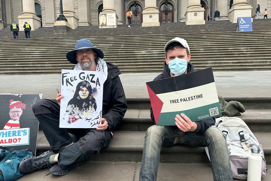 Two men sitting on steps, holding posters in support of ending the war in Gaza.