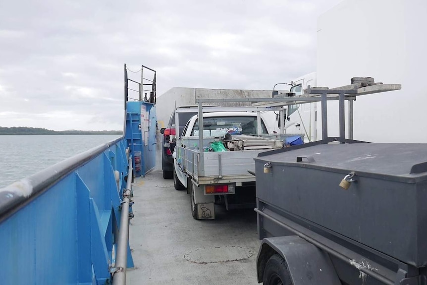 A barge on the water carries vehicles and a trailer, heading towards an island under cloudy skied.