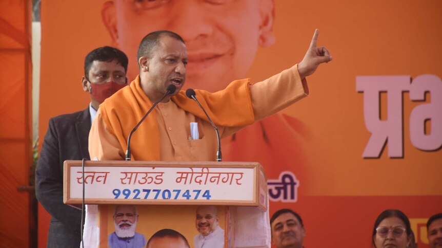 Yogi Adityanath giving speech on stage with one finger pointing out, wearing saffron robes against orange campaign background