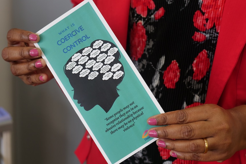 A blue pamphlet titled "What is Coercive Control" is held by a woman's hands. Her nails are manicured pink. 