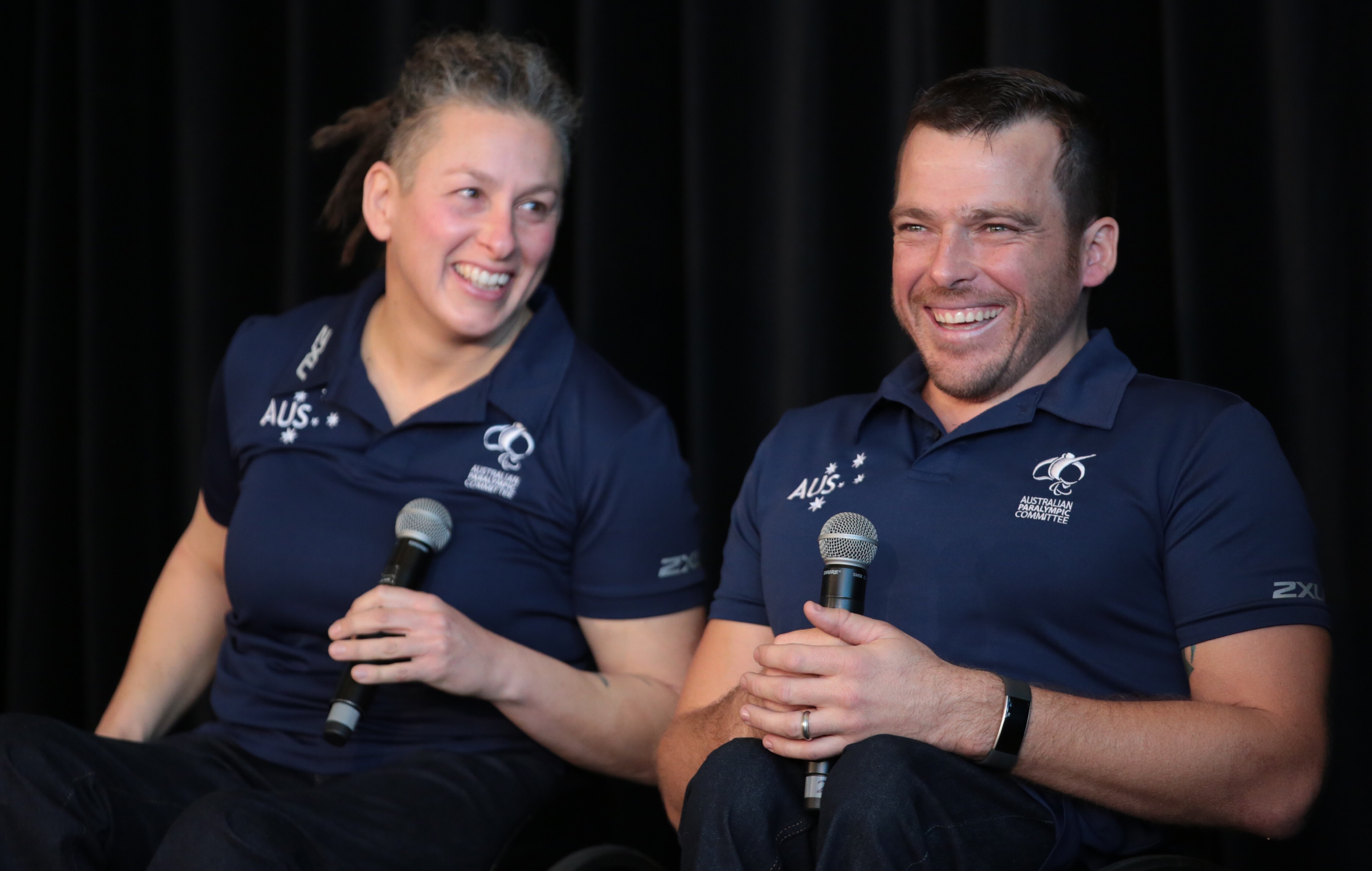 Two Australian Paralympians sit side by side at a press conference, laughing as they hold microphones.