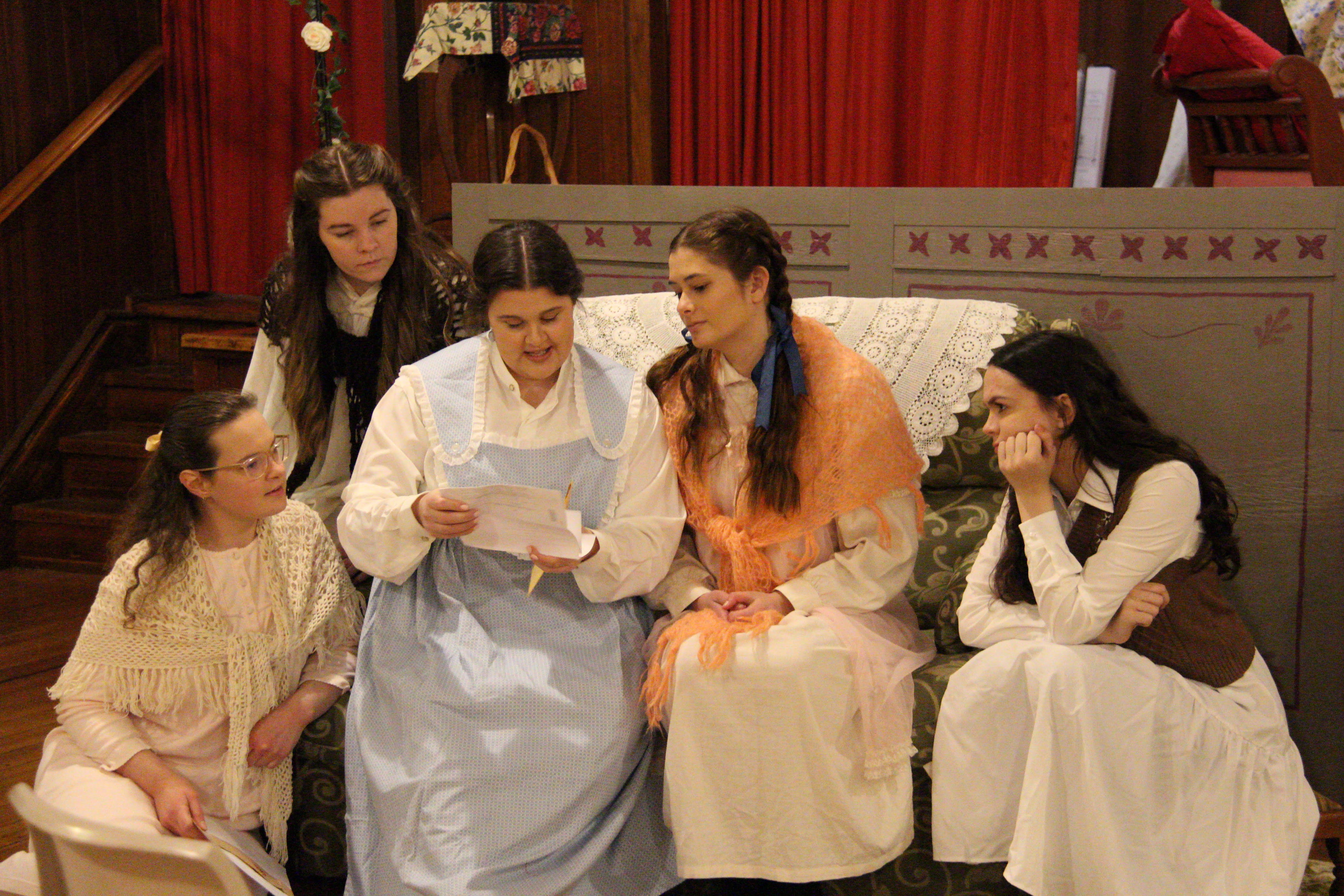 Four women sit and look at a letter. They are in costume for Little Women