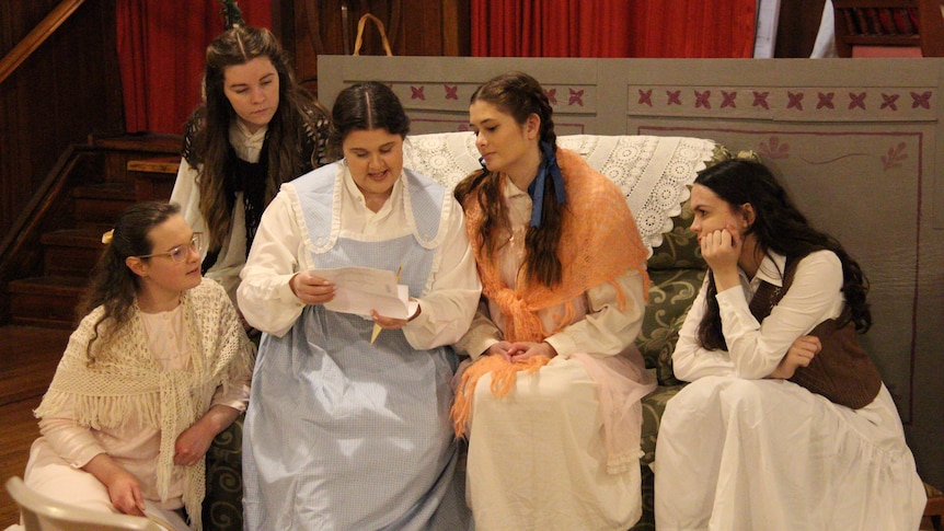Four women sit and look at a letter. They are in costume for Little Women