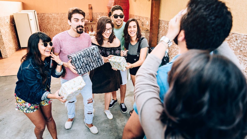 A group of people in a courtyard holding gifts ready to present gifts to a man who has his eyes shielded by a woman