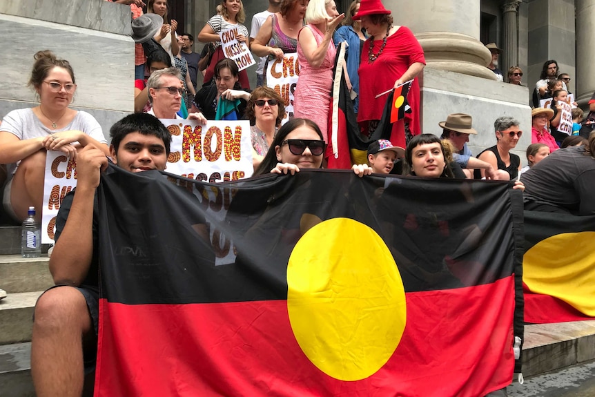Crowds with signs saying "C'MON AUSSIE C'MON" and holding Aboriginal flags protest on the steps of SA Parliament.