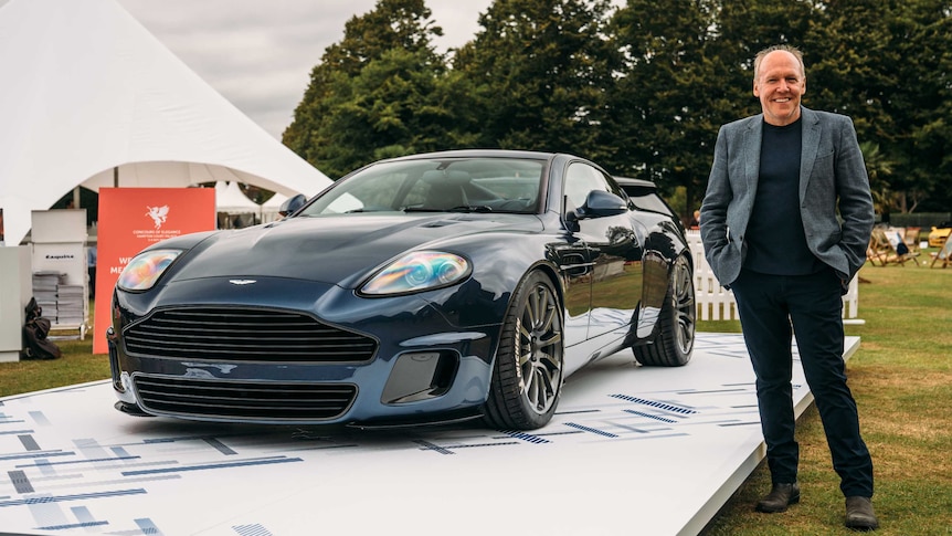 Ian Callum stands next to an Aston Martin Vanquish on a small white platform adjacent to a marquee in a green field.
