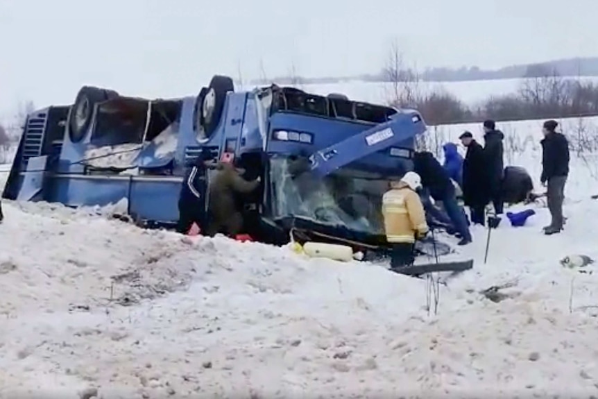 Emergency workers stand near a blue upturned bus in the snow.