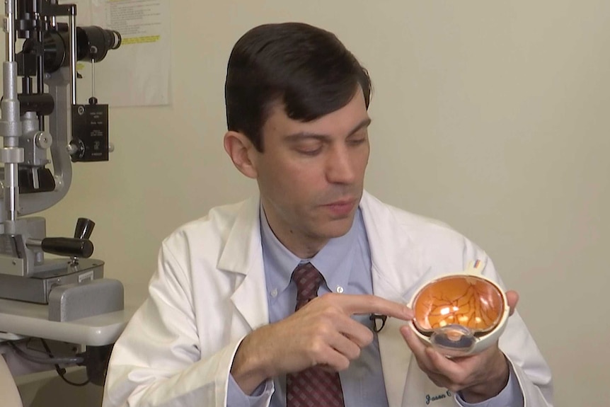 A man in a blue shirt and white doctor's coat points to a large, plastic model of an eye.