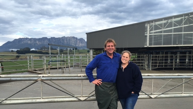 man and woman stand in front of milking sheds with mountain in background
