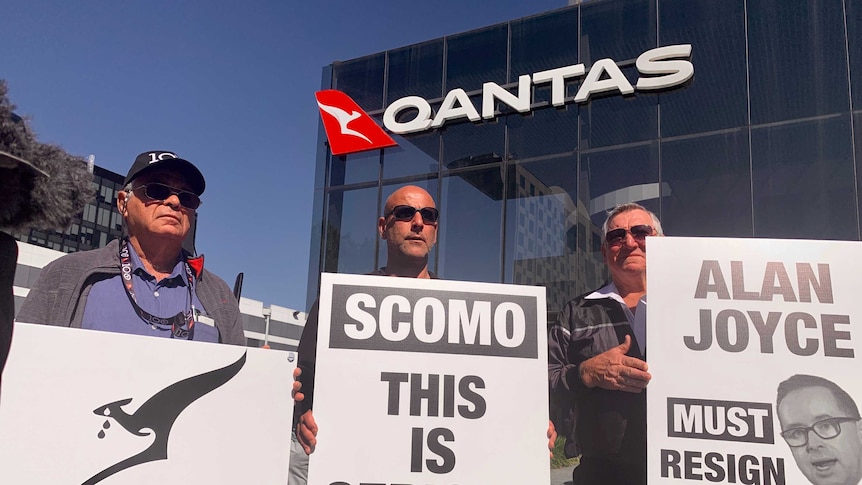 Three men hold signs protesting Qantas outsourcing, in front of the Qantas headquarters building.