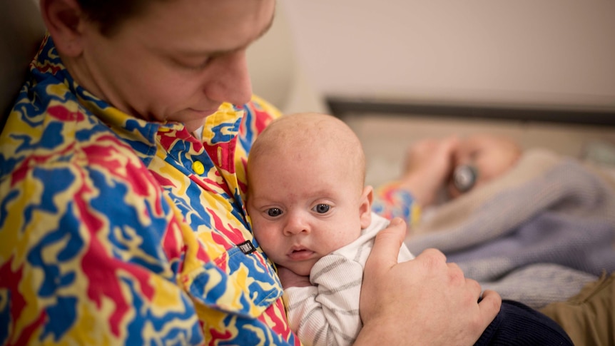 A man with a colourful shirt sitting on a couch with a baby