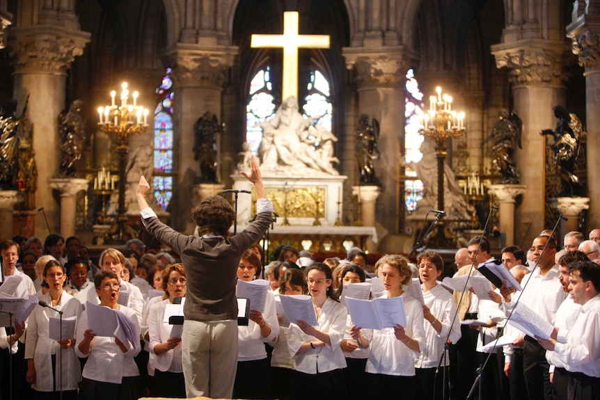 A conductor raises her hands as a group of choristers sing, candles burn and a cross is illuminated inside a cathedral.