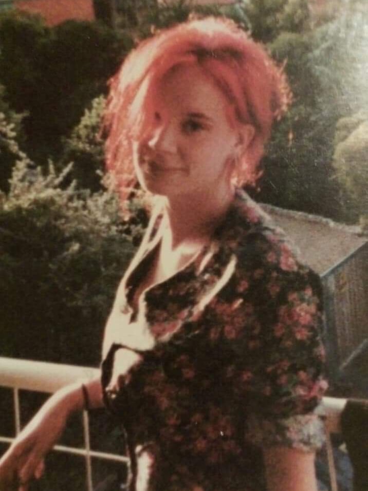 A young woman in a fuzzy photo smiles at the camera. She has pink hair