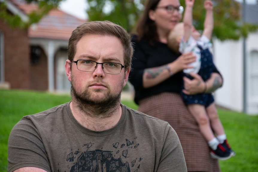 a man looking solemn to the camera, his wife and child in background