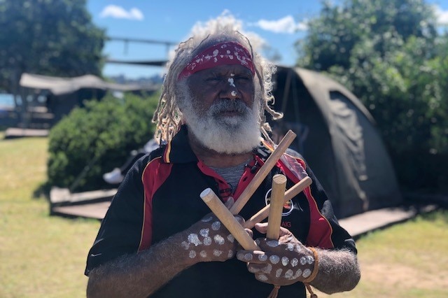 indigenous man in traditional skin markings and a red bandana holding percussion sticks