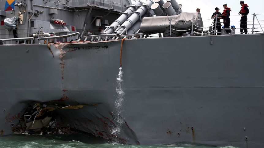 Sailors stand aboard a grey naval vessel that has a large dent in its side.