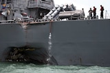 Sailors stand aboard a grey naval vessel that has a large dent in its side.