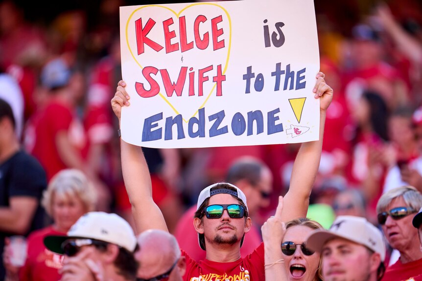 A man in sunglasses holds up a sign which reads 'Kelce is Swift to the Endzone'.