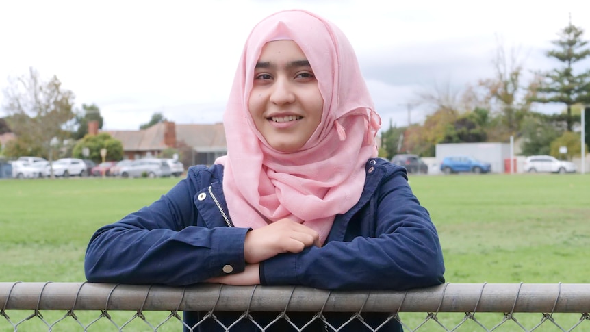 A close up shot of a teenage girl wearing a navy blue jacket and pink hijab leaning on a metal fence. 