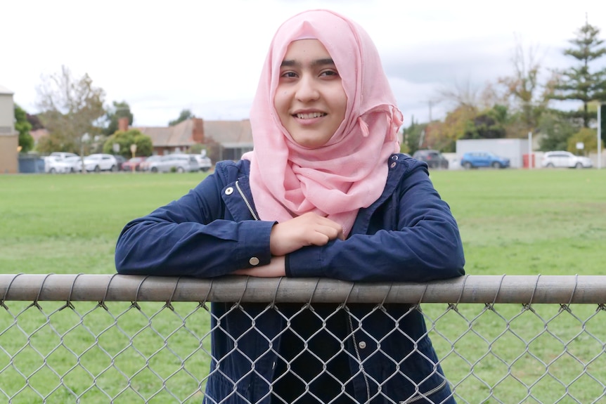 A close up shot of a teenage girl wearing a navy blue jacket and pink hijab leaning on a metal fence. 
