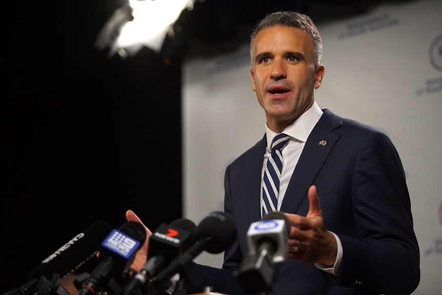 A man wearing a suit, white shirt and striped tie gestures with his hands in front of media microphones