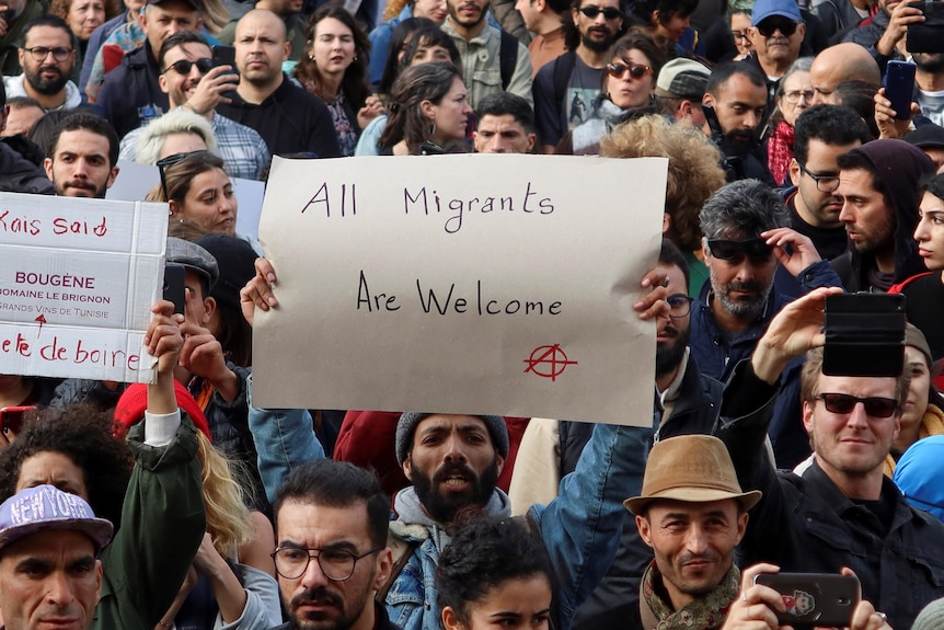 A man standing in a crowd of people holds up a sign saying 'All Migrants Are Welcome'