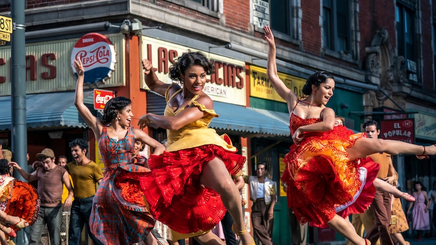 Three Puerto Rican women wearing vibrantly coloured dresses are mid high kick in a dance routine on the streets of New York.