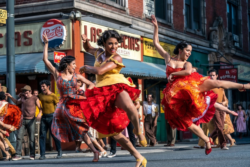 Three Puerto Rican women wearing vibrantly coloured dresses are mid high kick in a dance routine on the streets of New York.