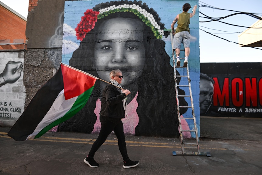 A person paints a mural of a young Palestinian girl on a wall with a woman holding a Palestine flag walking in the foreground