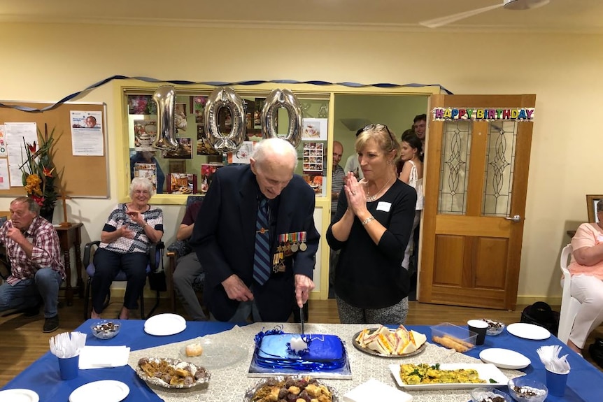 Mr Willing holding a knife and cutting a cake at his 100th birthday with a rom full of other people