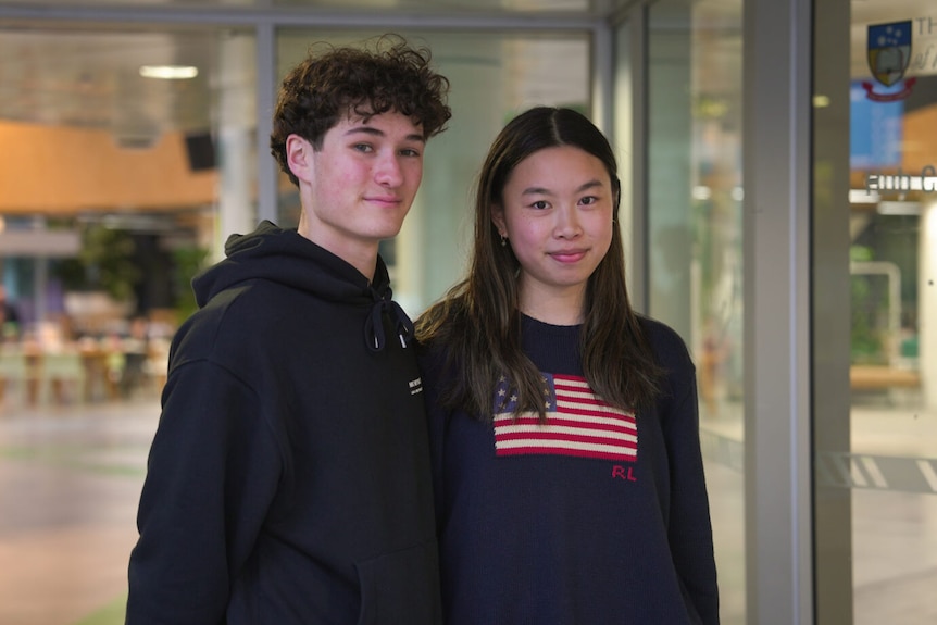 A teenage girl and boy stand next to each other in a university building, smiling.