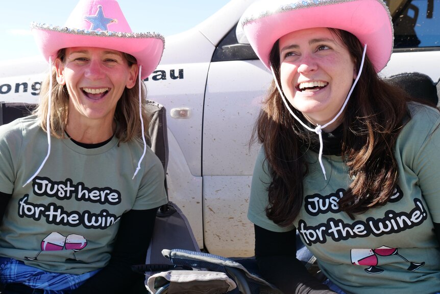 Two women wearing pink cowboy hats sitting infront of a car in camping chairs smiling. 