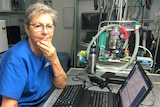 A woman with grey hair and glasses, dressed in scrubs, sits in an office in front of a computer.