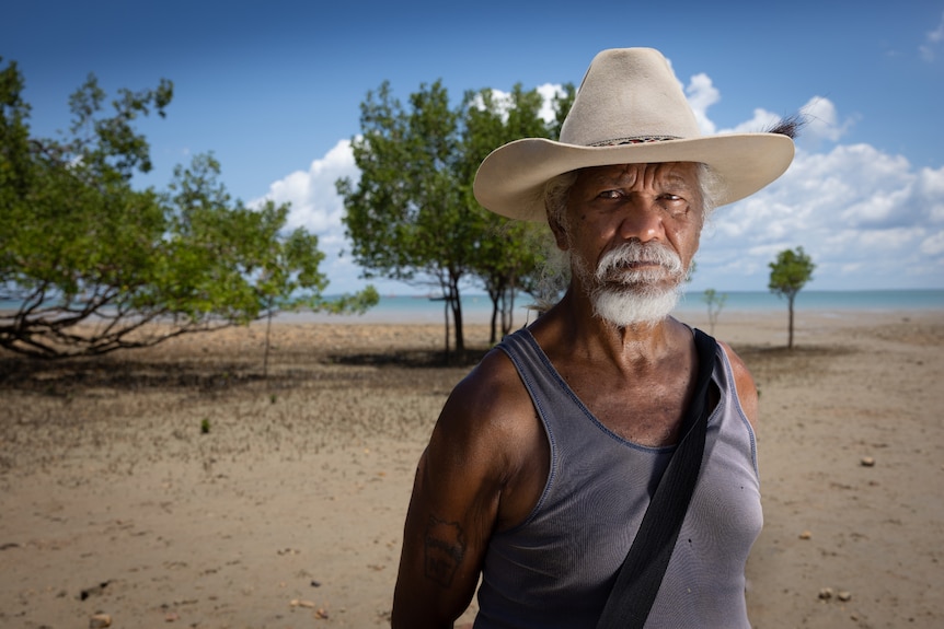 A serious-looking man in a cowboy hat standing on a beach with mangroves and the ocean in the background.