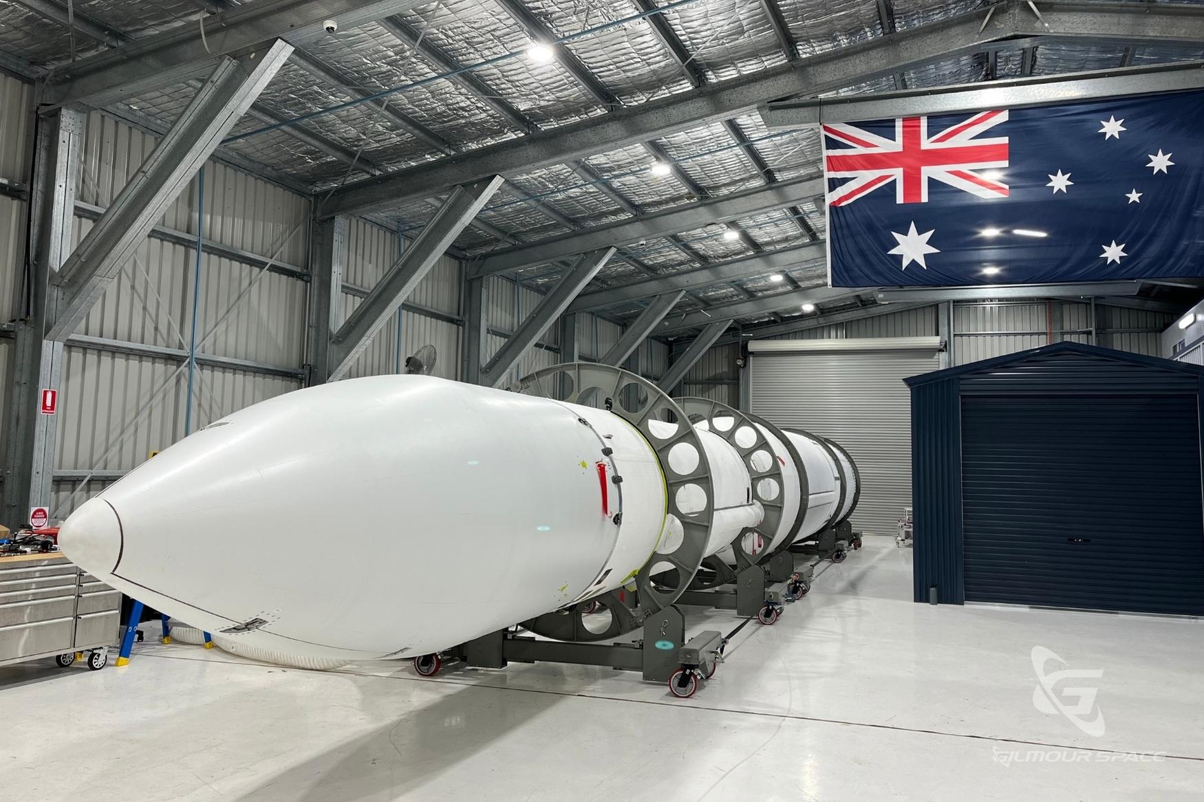 White rocket sits in hangar with Australian flag in background.
