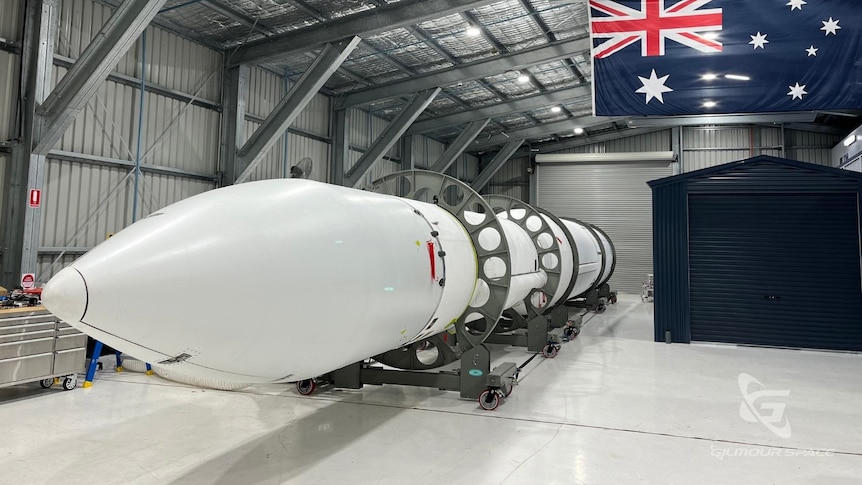 White rocket sits in hangar with Australian flag in background.