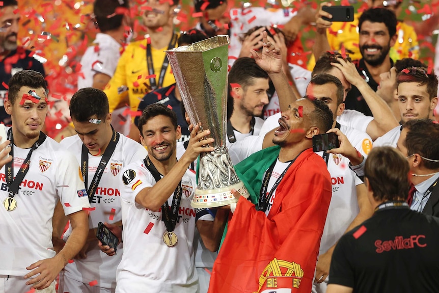 As confetti falls, Sevilla players smile and celebrate with the trophy. One player is wearing a Portuguese flag on his shoulders