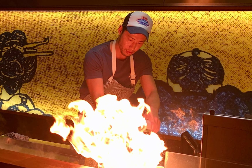 A chef in a cap and apron fires up the teppanyaki grill in a Japanese restaurant, with flames leaping upwards.