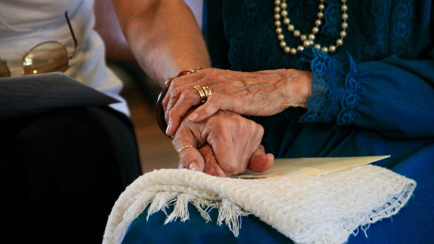 An elderly woman holds onto a younger person's arm while sitting together.