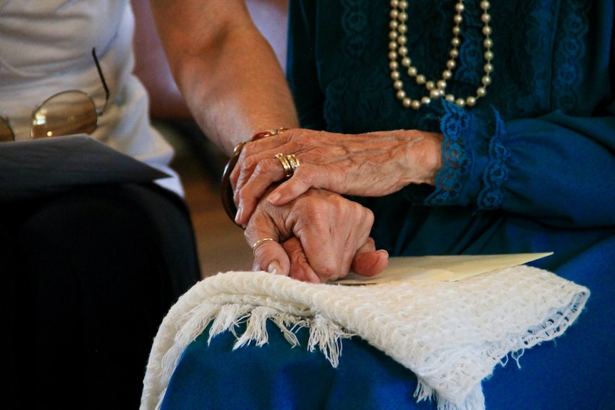 An elderly woman holds onto a younger person's arm while sitting together.