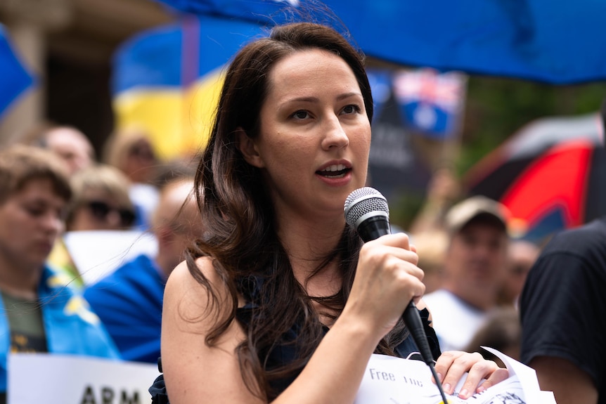 A young woman speaks into a microphone at a rally with Ukrainian and Australian flags visible in the background